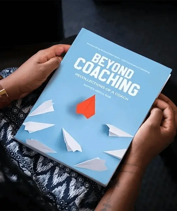 Beyond Coaching: Recollections of a Coach — light blue paperback with paper airplanes held in a reader’s hands